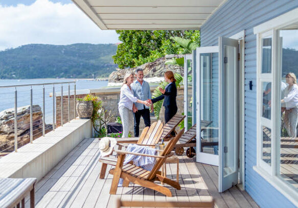 Real estate agent showing a mature couple a new house. The house is contemporary. All are happy and smiling and shaking hands. The couple are casually dressed and the agent is in a suit. Waterfront can be seen in the background
