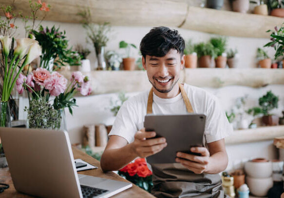 Asian male florist, owner of small business flower shop, using digital tablet while working on laptop against flowers and plants. Checking stocks, taking customer orders, selling products online. Daily routine of running a small business with technology